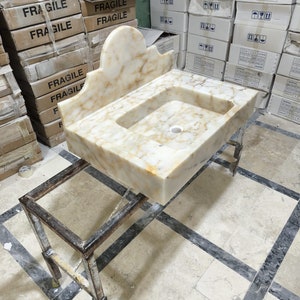 Puede incluir: Un lavabo rectangular de m&aacute;rmol con respaldo decorativo, con un patr&oacute;n veteado beige claro y marr&oacute;n. El lavabo est&aacute; sostenido por un soporte met&aacute;lico. En el fondo hay cajas etiquetadas como "FR&Aacute;GIL".