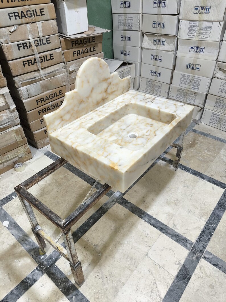 Puede incluir: Un lavabo rectangular de m&aacute;rmol con respaldo decorativo, con un patr&oacute;n veteado beige claro y marr&oacute;n. El lavabo est&aacute; sostenido por un soporte met&aacute;lico. En el fondo hay cajas etiquetadas como "FR&Aacute;GIL".