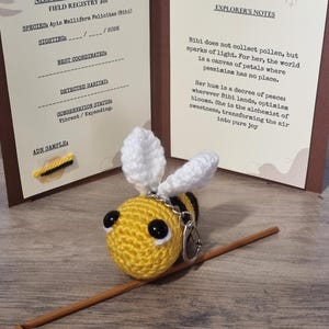 May include: A crocheted bee keychain with yellow, black, and white details. The bee has large black eyes and a silver-colored keychain ring. The background includes two cards with text, including "Explorer's Notes" and "Field Registry".
