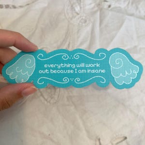 May include: A turquoise bookmark with a scalloped edge and white line art featuring two stylized wings on the sides. The text in the center reads, everything will work out because I am insane, written in a white, pixelated, cross-stitch style font.