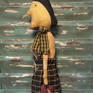 May include: A handmade rag doll with a black and yellow plaid skirt, a black and yellow checkered scarf, and a brown house-shaped object in her hand. The doll is hanging on a weathered blue wooden surface.