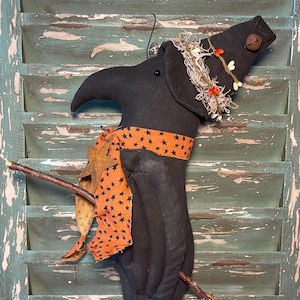May include: A black Halloween decoration shaped like a witch, with a pointed hat and a star-patterned orange scarf. The witch has a twig broom and is adorned with a bell and a decorative garland. The background is a distressed, teal-colored shutter.