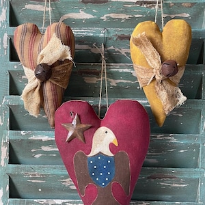 May include: Three heart-shaped fabric ornaments hanging by strings. One is striped brown and tan, one is solid mustard yellow, and one is deep red featuring a bald eagle graphic and a small metal star. Each has a burlap bow and a metal bell.