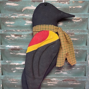 May include: A black bird-shaped stuffed toy with a red, yellow, and black wing and a yellow and green plaid scarf. The toy is hanging on a weathered green wooden surface.