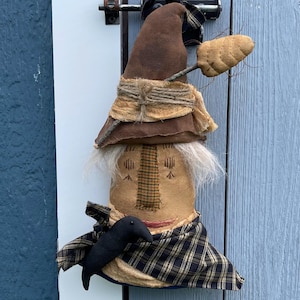 May include: A handmade, folk art doll with a pointed brown hat and a plaid scarf. The doll has a tan face with stitched features, white hair, and a small black bird attached. A decorative piece for the home.