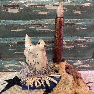 May include: A rustic decorative arrangement featuring a fabric bird with black polka dots, a faux candle, and straw accents. The candle has a brown, textured base and a cream-colored tip. The arrangement sits on a blue base with a burlap accent.