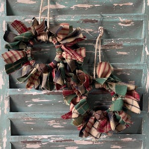 May include: Two small, handmade fabric wreaths with plaid patterns in red, green, and navy blue. Each wreath is adorned with fabric strips and a small bell. They hang from twine on a distressed, turquoise-painted wooden shutter.