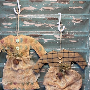 May include: Two handmade fabric ornaments in the shape of people, hanging from hooks on a weathered blue wooden surface. The ornament on the left is wearing a brown and white patterned dress with a burlap skirt and a brown bell. The ornament on the right is wearing a brown and black plaid dress with a burlap skirt and a brown bell.