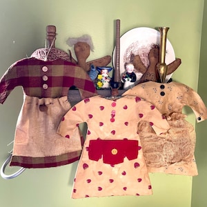 May include: Three handmade rag dolls dressed in vintage-inspired clothing. The dolls are wearing a plaid dress, a strawberry print dress, and a floral print dress. The dolls are displayed on a shelf with other decorative items.
