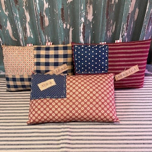 May include: Four handmade fabric pillows with red, white, and blue patchwork designs. Each pillow has a different patriotic word on it: "USA", "Glory", "America", and a star pattern. The pillows are on a striped fabric surface.