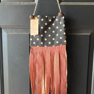 May include: A patriotic wall hanging with a black top adorned with gold stars and a red and tan striped bottom. The tag reads "GOD BLESS THE USA". The hanging is suspended by a twine loop.