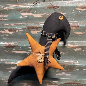 May include: A handmade black fabric bird with an orange star body, adorned with a button and twine. The bird wears a black and tan scarf. A decorative piece for seasonal or rustic decor.