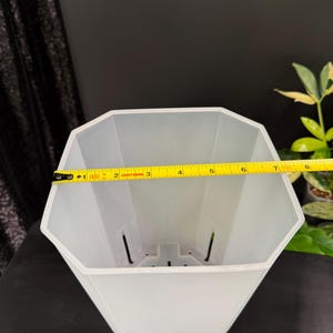 May include: A white, geometric-shaped plant pot with a measuring tape showing a width of approximately 7 inches. The pot has drainage holes and a textured interior. The background is dark, with a glimpse of a plant.