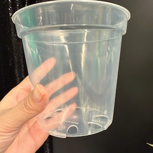 May include: A clear plastic plant pot with a wide rim and drainage holes. The pot is held up by a hand. The pot is designed for planting and gardening.