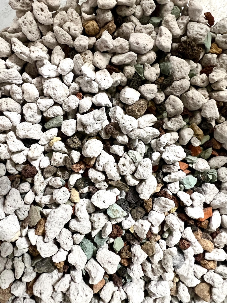 May include: A collection of small, porous volcanic rocks in various shades of white, light gray, dark brown, and pale green. The stones have an irregular, jagged texture with visible holes and crevices, creating a dense, multi-colored aggregate.