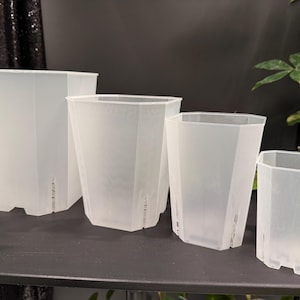 May include: Four clear, plastic plant pots of varying sizes are arranged on a dark surface. The pots have a geometric, faceted design and drainage holes near the base. The pots are empty and ready for planting.
