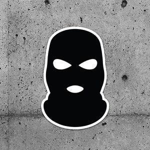 May include: Two black and white ski mask stickers. The stickers are black with white eye and mouth openings, and a white border. The stickers are displayed against a gray concrete background.