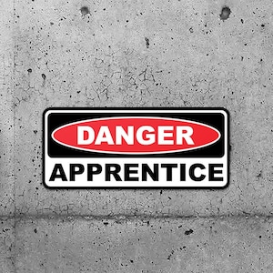 May include: Two black and red warning signs with the text "DANGER APPRENTICE" on a grey concrete wall.