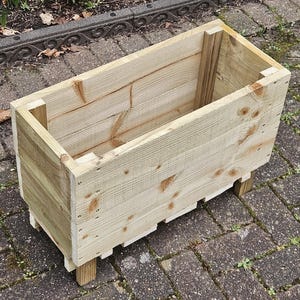 May include: A wooden planter box with a natural finish. The planter is made of unfinished wood planks and has a rectangular shape. The planter is sitting on a brick patio.