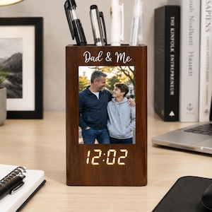 Könnte beinhalten: Ein Holz-Stiftehalter mit einem Foto von Vater und Sohn, dem Schriftzug "Dad & Me" und einer Digitaluhr, die 12:02 anzeigt. Der Halter enthält Stifte und steht auf einem Schreibtisch mit Büchern und einem Laptop.
