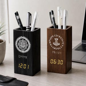 May include: Two rectangular wooden pen holders with digital clocks. The black holder features a white wolf graphic and runes, displaying 12:01. The brown holder features a white Mjolnir hammer graphic and runes, displaying 06:30. Both hold pens.