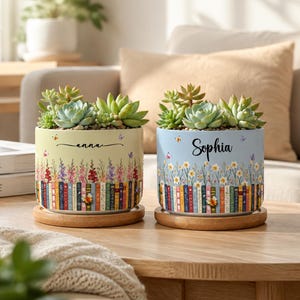May include: Two cylindrical ceramic planters with wooden saucers, each containing green succulents. The left planter is pale yellow with the name Anna, while the right is light blue with the name Sophia. Both feature floral and book spine prints.