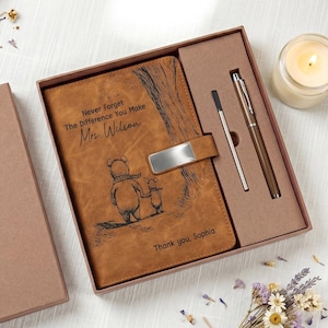 May include: A brown leather journal and pen set in a gift box. The journal has an engraved Winnie the Pooh illustration and the text "Never Forget The Difference You Make, Mrs. Wilson". A silver pen and a black pen are included.