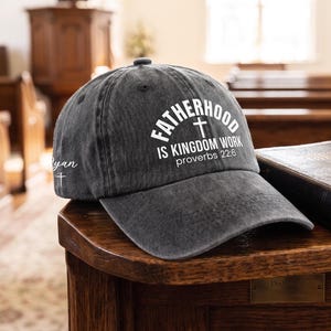 May include: A charcoal gray baseball cap with the text "FATHERHOOD IS KINGDOM WORK" and a cross graphic. The cap also features the text "proverbs 22:6". The hat is sitting on a wooden surface.