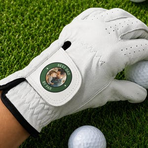 K&ouml;nnte beinhalten: Ein wei&szlig;er Golfhandschuh aus Leder mit einem kreisf&ouml;rmigen Aufn&auml;her am Handgelenkriemen. Der Aufn&auml;her ist dunkelgr&uuml;n mit wei&szlig;em Rand und zeigt ein Foto einer Person, die ein Kind h&auml;lt, mit dem in Wei&szlig; gedruckten Text BEST DAD BY PAR.