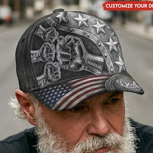 May include: A black baseball cap with a patriotic design. The cap features an American flag on the brim and a graphic of fists and stars. The names "Tim", "Abby", "Jake", and "Charlie" are printed on the cap. The text "Dad" is also visible.