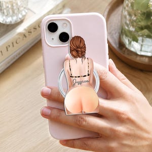 Pode incluir: Uma capa de smartphone rosa claro com um suporte circular. O suporte exibe a ilustração de uma pessoa com cabelo castanho em um coque, vestindo um top preto de alças com o nome Jessica impresso em branco na parte das costas.