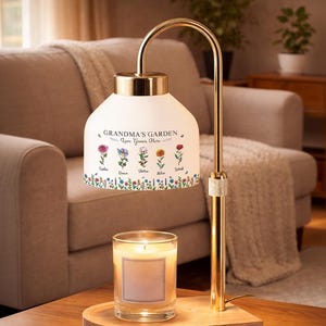 May include: A gold-toned lamp with a white shade featuring the text "Grandma's Garden" and floral illustrations. A lit candle sits on a wooden base below the lamp. The lamp is on a wooden table.