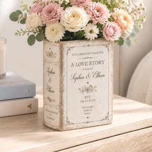 May include: A rectangular ceramic vase shaped like a book, featuring the text "OUR GREATEST CHAPTER A LOVE STORY written by Sophia & Ethan EST. 04.12.2025". The vase is filled with a bouquet of pink, white, and cream flowers and green foliage.