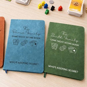 May include: Three rectangular game night score books in tan, blue, and green with a textured finish. Each cover features a family name, the text GAME NIGHT SCORE BOOK, illustrations of dice, dominoes, and cards, and the text WHO'S KEEPING SCORE?