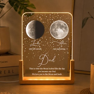 May include: A personalized night light with a wooden base and a clear acrylic panel. The panel features two moon phases, names, and dates, with the word "Dad" and a heartfelt message. The light glows with a warm, inviting hue.