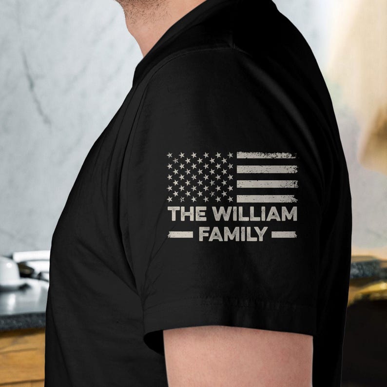 May include: Black t-shirt featuring a distressed American flag design on the upper sleeve. Below the flag, the text reads "THE WILLIAM FAMILY" in a white, sans-serif font, with a line above and below the text.