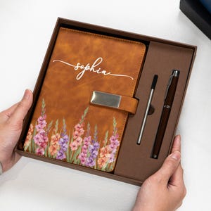 Puede incluir: Un diario de cuero marr&oacute;n con un dise&ntilde;o floral y el nombre "Sophia" en escritura blanca. El diario est&aacute; en una caja de regalo con un bol&iacute;grafo y un l&aacute;piz &oacute;ptico. El diario tiene un cierre plateado.