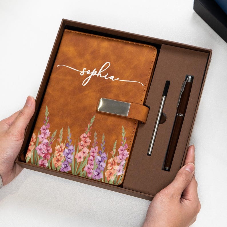 Puede incluir: Un diario de cuero marr&oacute;n con un dise&ntilde;o floral y el nombre "Sophia" en escritura blanca. El diario est&aacute; en una caja de regalo con un bol&iacute;grafo y un l&aacute;piz &oacute;ptico. El diario tiene un cierre plateado.