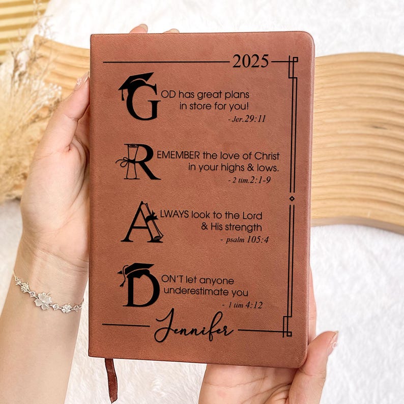 May include: A brown leather journal with the word "GRAD" in large letters. Each letter has a graduation cap and scroll design. The journal has inspirational quotes and bible verses for graduates. The year 2025 and the name "Jennifer" are also printed on the cover.
