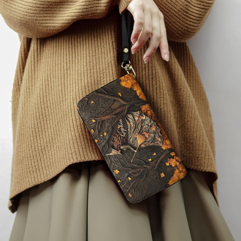 May include: A dark brown clutch with a detailed autumn-themed design featuring trees, leaves, and the name "Sophia" in gold lettering. The clutch has a black wrist strap and gold-colored hardware.