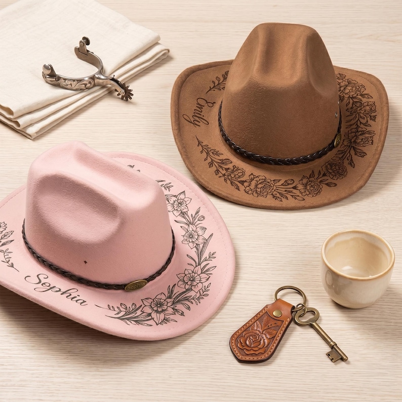 Pode incluir: Um chap&eacute;u de cowboy rosa claro com uma faixa tran&ccedil;ada escura e gravuras florais com o nome Sophia, e um chap&eacute;u de cowboy bege com uma faixa tran&ccedil;ada escura e gravuras de rosas com o nome Emily, junto a uma espora e um porta-chaves.