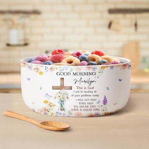 Op de afbeelding: Een witte keramische kom gevuld met kleurrijke ontbijtgranen en fruit. De kom is versierd met bloemmotieven, vlinders en de tekst "GOOD MORNING Marilyn". Een houten lepel ligt op de tafel.