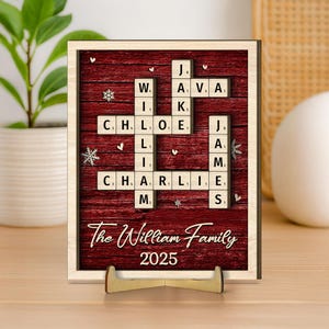 May include: A personalized wooden family name sign with a red background and a light wood frame. The sign features a crossword puzzle design with family names and the year 2025. Decorative snowflakes and hearts are scattered across the surface.