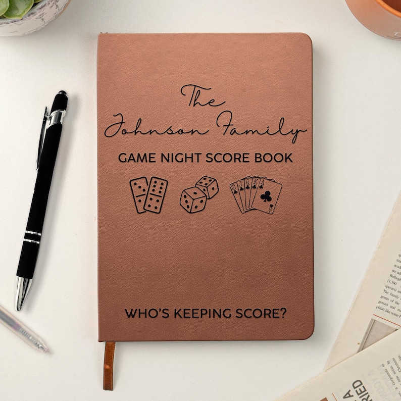 May include: A copper-colored notebook with black text reading The Johnson Family GAME NIGHT SCORE BOOK and WHO'S KEEPING SCORE? The cover features black line art of a domino, two dice, and a hand of playing cards, accompanied by a black pen.