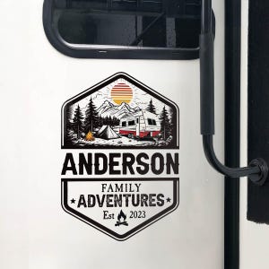 Può includere: Un adesivo in bianco e nero raffigurante una scena di campeggio con una roulotte, una tenda, montagne e alberi. L'adesivo include il nome "ANDERSON" e il testo "FAMILY ADVENTURES Est 2023".