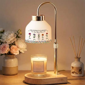 May include: A decorative lamp with a white shade featuring the text "GRANDMA'S GARDEN Love Grows Here" and floral illustrations. The lamp has a gold-colored metal frame and sits on a wooden base. A lit candle and a vase with flowers are nearby.