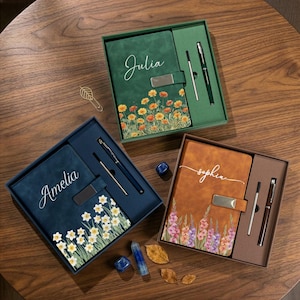 Puede incluir: Tres juegos de diario y bol&iacute;grafo personalizados en cajas de regalo. Los diarios son verdes, azules y marrones, cada uno con un dise&ntilde;o floral y un nombre en cursiva. Cada juego incluye un bol&iacute;grafo a juego.