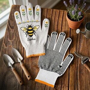 Op de afbeelding: Een paar witte tuinhandschoenen met een gele en zwarte bijenillustratie, kleine gele bloempatronen en de naam Sophia in zwarte schrijfletters. E&eacute;n handschoen heeft een zwart stippenpatroon op de handpalm en de vingers voor extra grip.