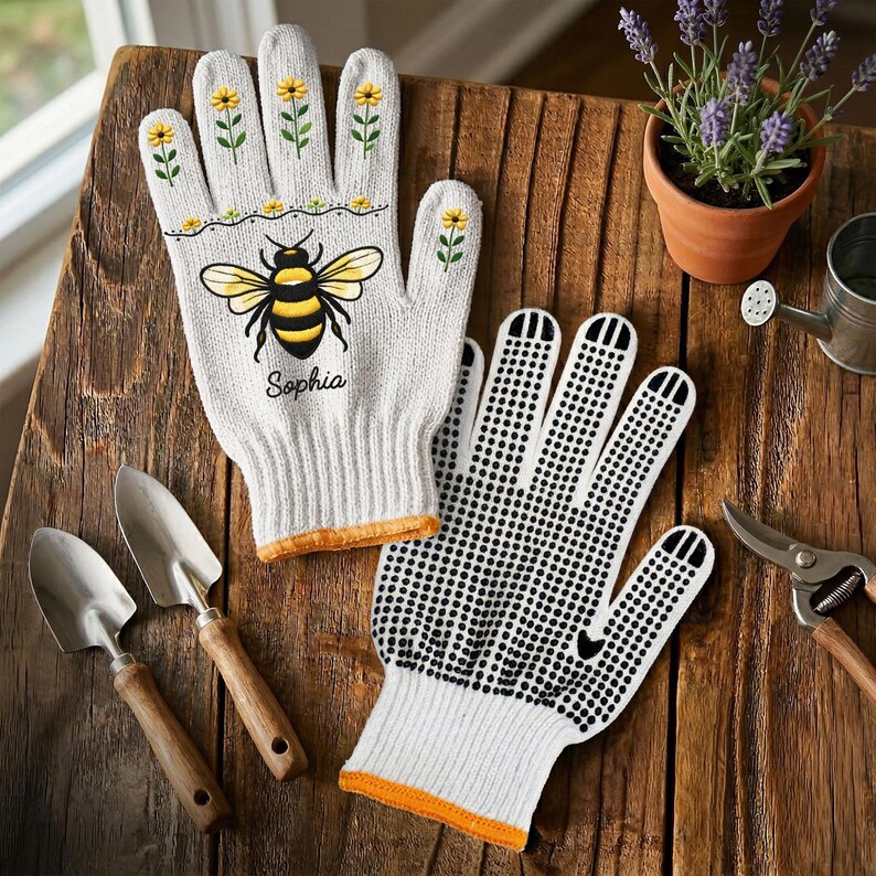 Op de afbeelding: Een paar witte tuinhandschoenen met een gele en zwarte bijenillustratie, kleine gele bloempatronen en de naam Sophia in zwarte schrijfletters. E&eacute;n handschoen heeft een zwart stippenpatroon op de handpalm en de vingers voor extra grip.