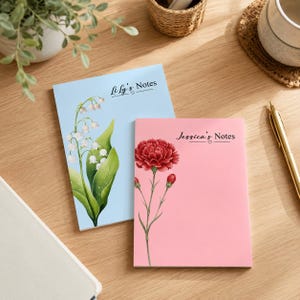 Peut inclure: Deux blocs-notes rectangulaires avec des illustrations florales. Le bloc-notes bleu clair présente un muguet et le texte "Lily's Notes". Le bloc-notes rose présente un œillet rouge et le texte "Jessica's Notes".